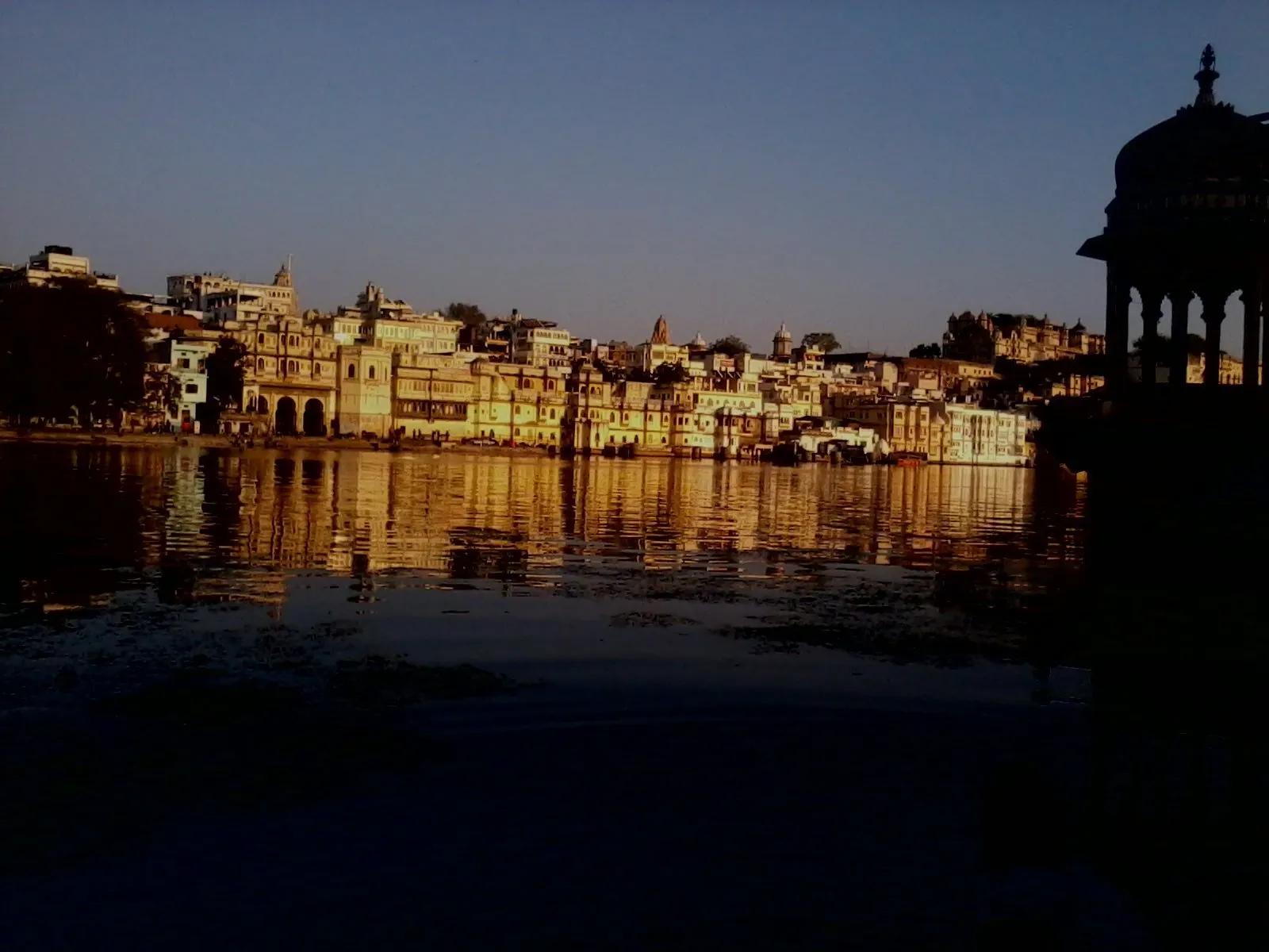 Golden-hour view across Lake Pichola of the old-city haveli row in Udaipur, cream buildings reflected in calm water with a domed chhatri to the right.