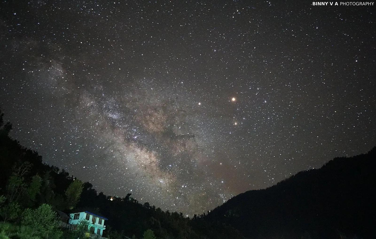 Milky Way galactic core rising over a silhouetted Himalayan ridge at night, with a single lit mountain home in the foreground