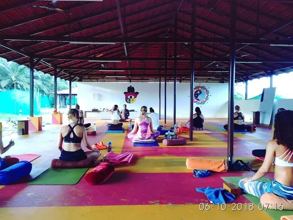 A yoga teacher training class in an open thatched shala in Goa, about ten students and a teacher seated cross-legged on colourful mats, palm grove behind
