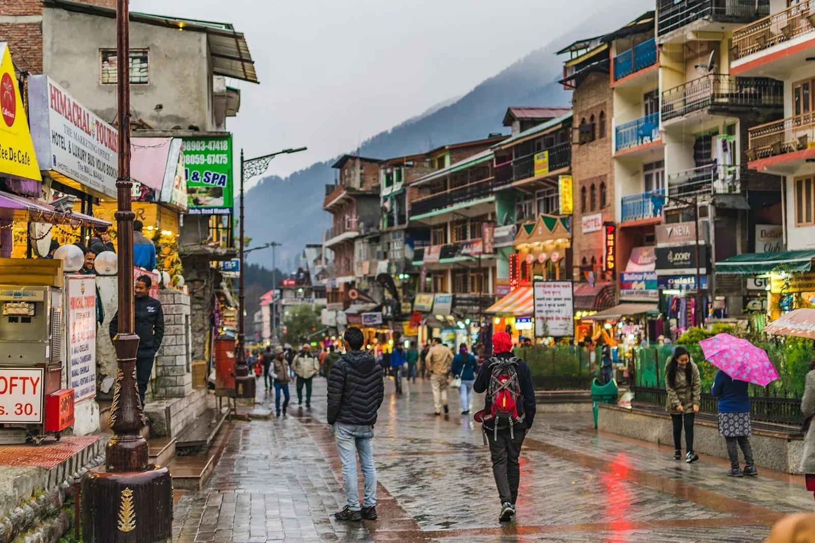 Tourists with umbrellas walking down Mall Road, Manali, on a rainy afternoon, pastel shop signs in Hindi and English