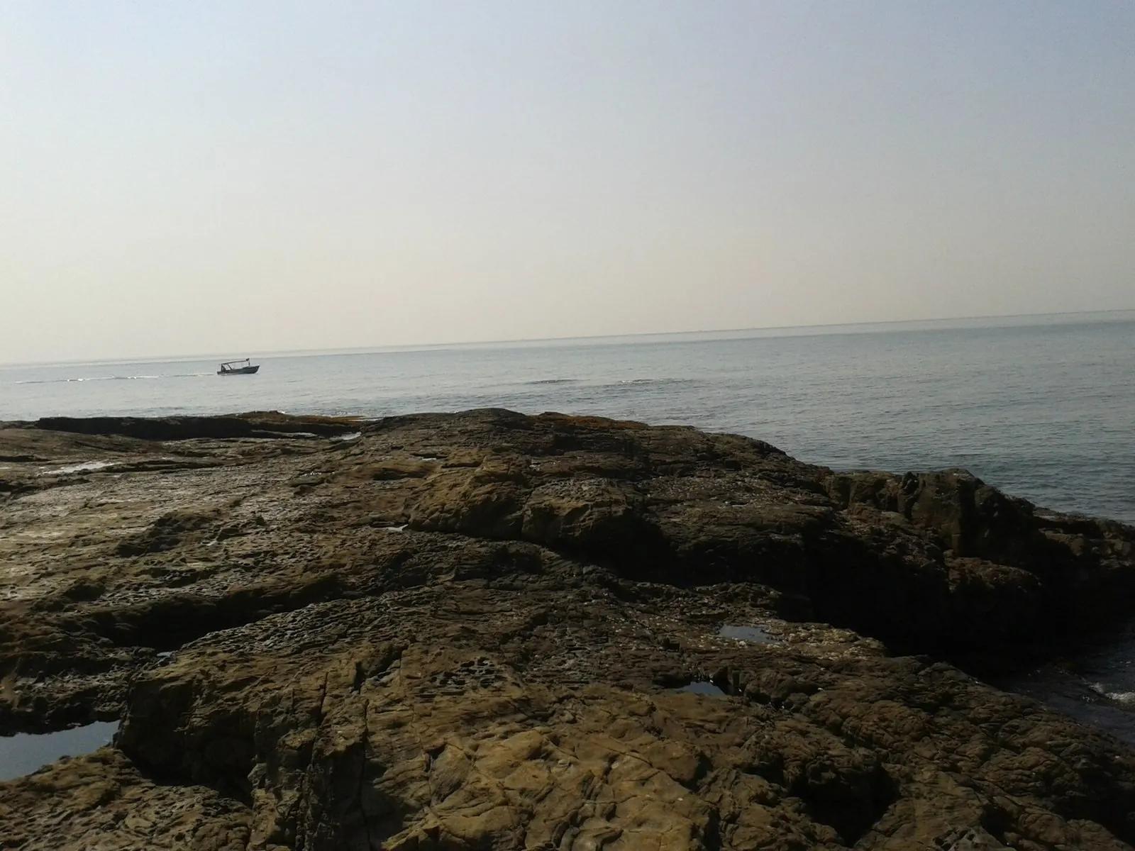 A flat dark rocky intertidal shelf at Anjuna beach, Goa, with small water-filled pools in the foreground rock and the calm sea and a fishing boat beyond under a pale sky.