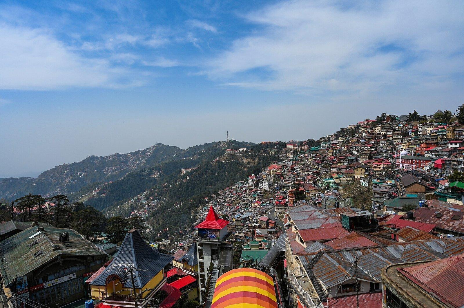 Shimla hillside cityscape with colourful buildings climbing the slope, mountains behind under clear sky