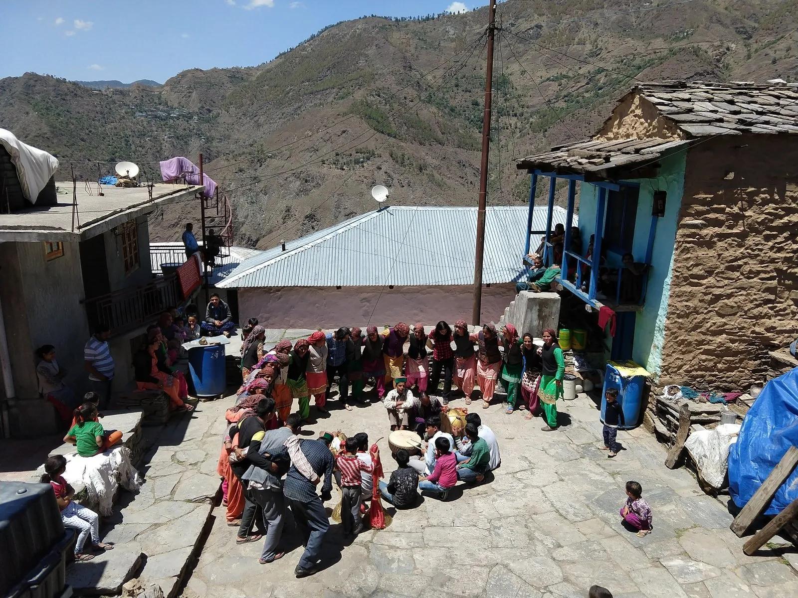 Women in traditional Kullu pattu dress and dhattu caps standing in a Nati formation in a village courtyard, seated audience in front, mountains and stone-roofed houses behind