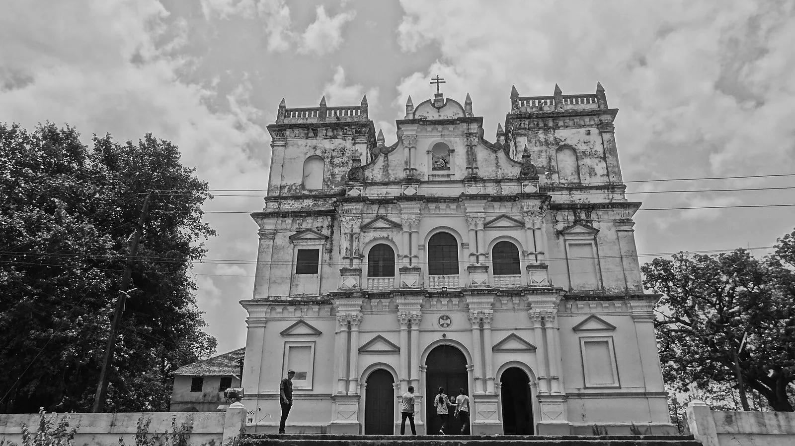 The Baroque facade of the Church of Our Lady of Compassion on the hill in Piedade, Divar Island, in black and white, a cross on the pediment and a few figures at the door