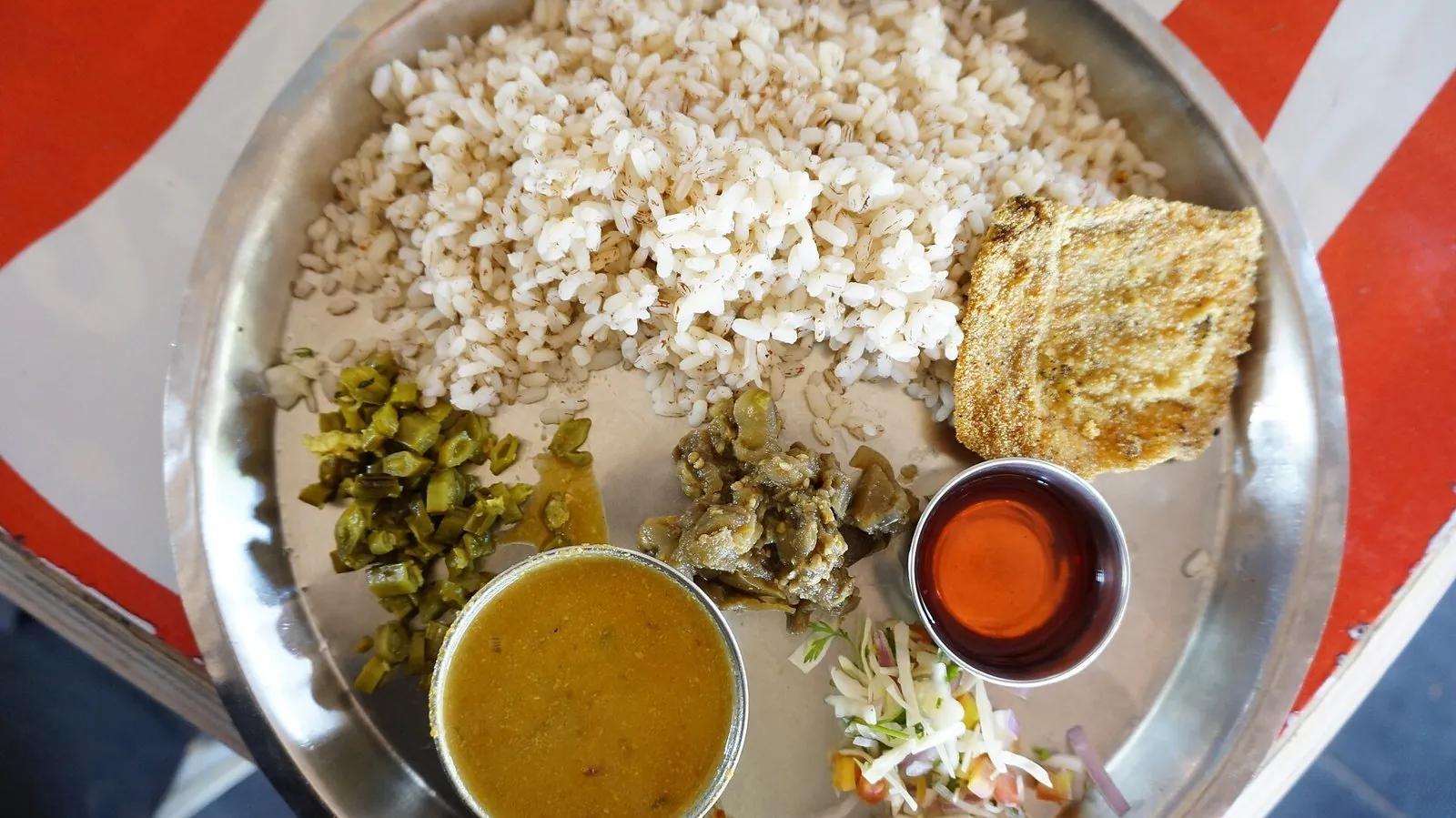A Goan fish thali on a steel plate: a mound of red parboiled rice, a bowl of fish curry, a piece of fried mackerel, pickled vegetables and a salad