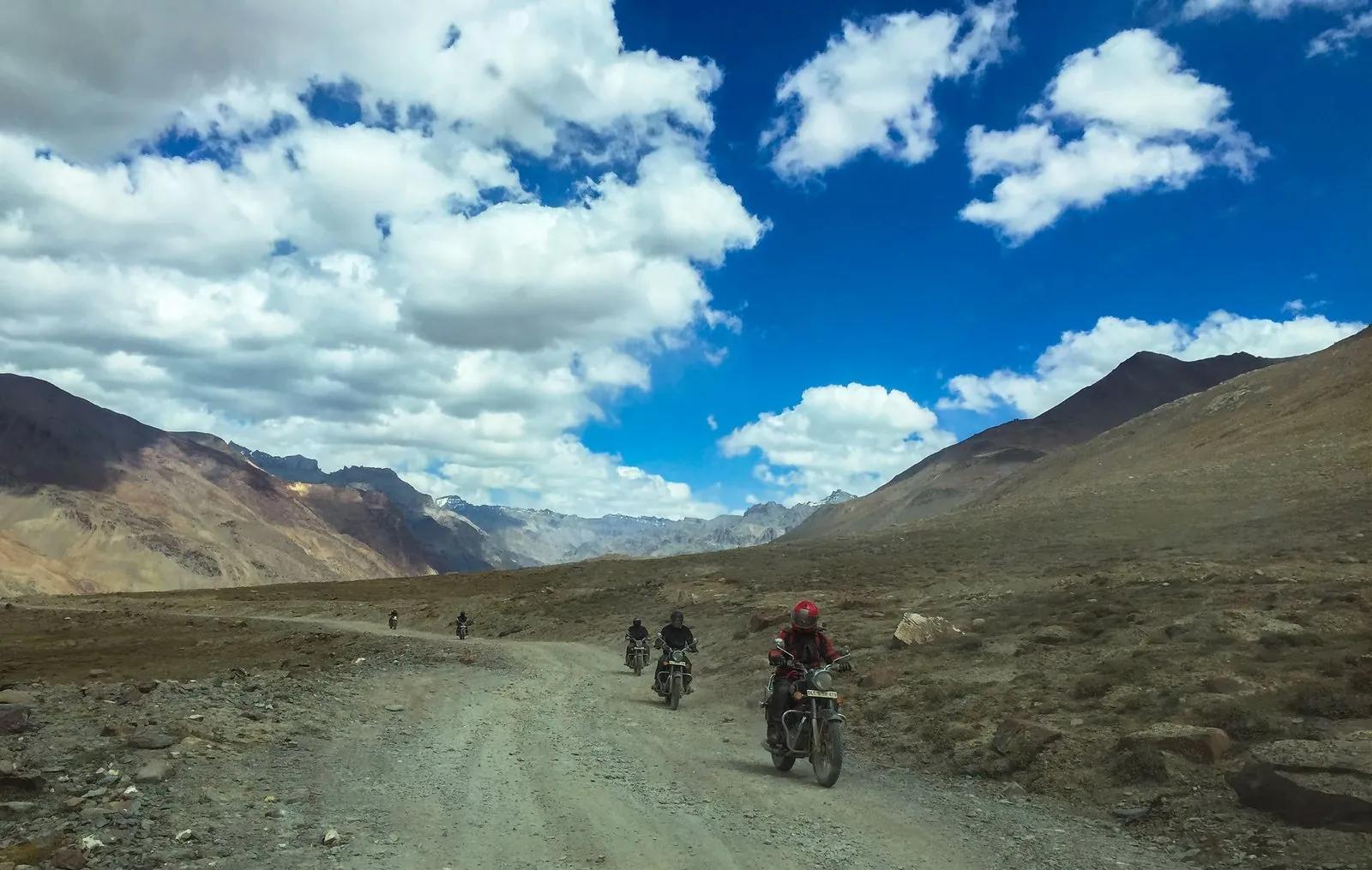 Four motorcyclists in red and black riding gear on a dusty unpaved Spiti Valley road with brown barren mountains rising on both sides and dramatic cloud formations overhead