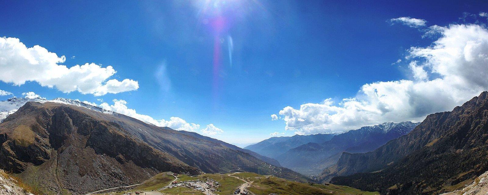 Wide panoramic view of the Manali valley from the Rohtang approach road, with the Kullu valley stretching into the distance under blue sky and white clouds