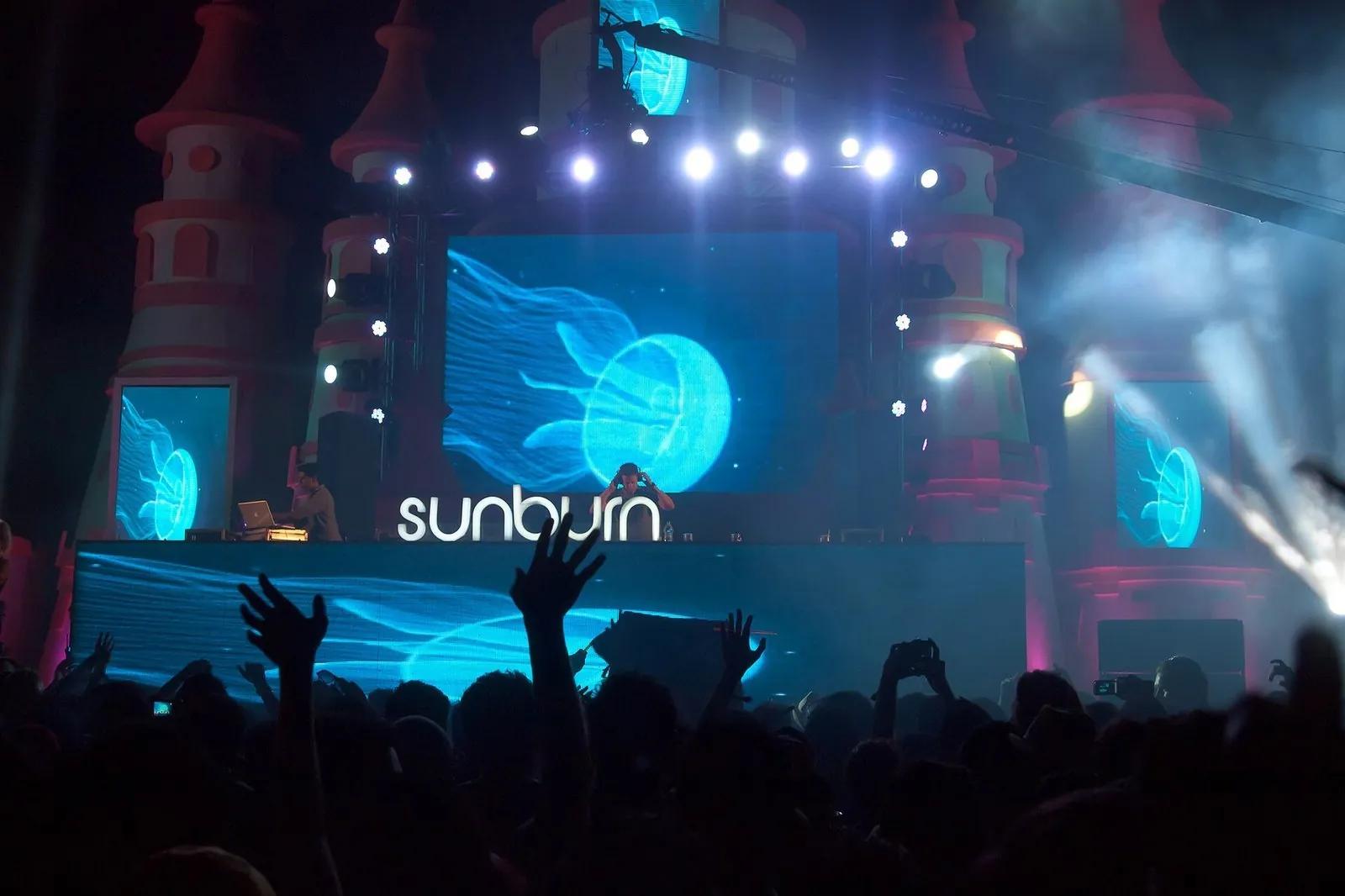 A huge EDM festival stage at night with a castle set, a DJ, blue jellyfish visuals and a crowd with hands raised, the word Sunburn lit up