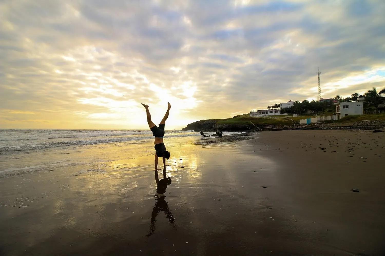 A person in silhouette holding a handstand on a wet beach at sunset, reflected in the sand, a palm-lined headland behind