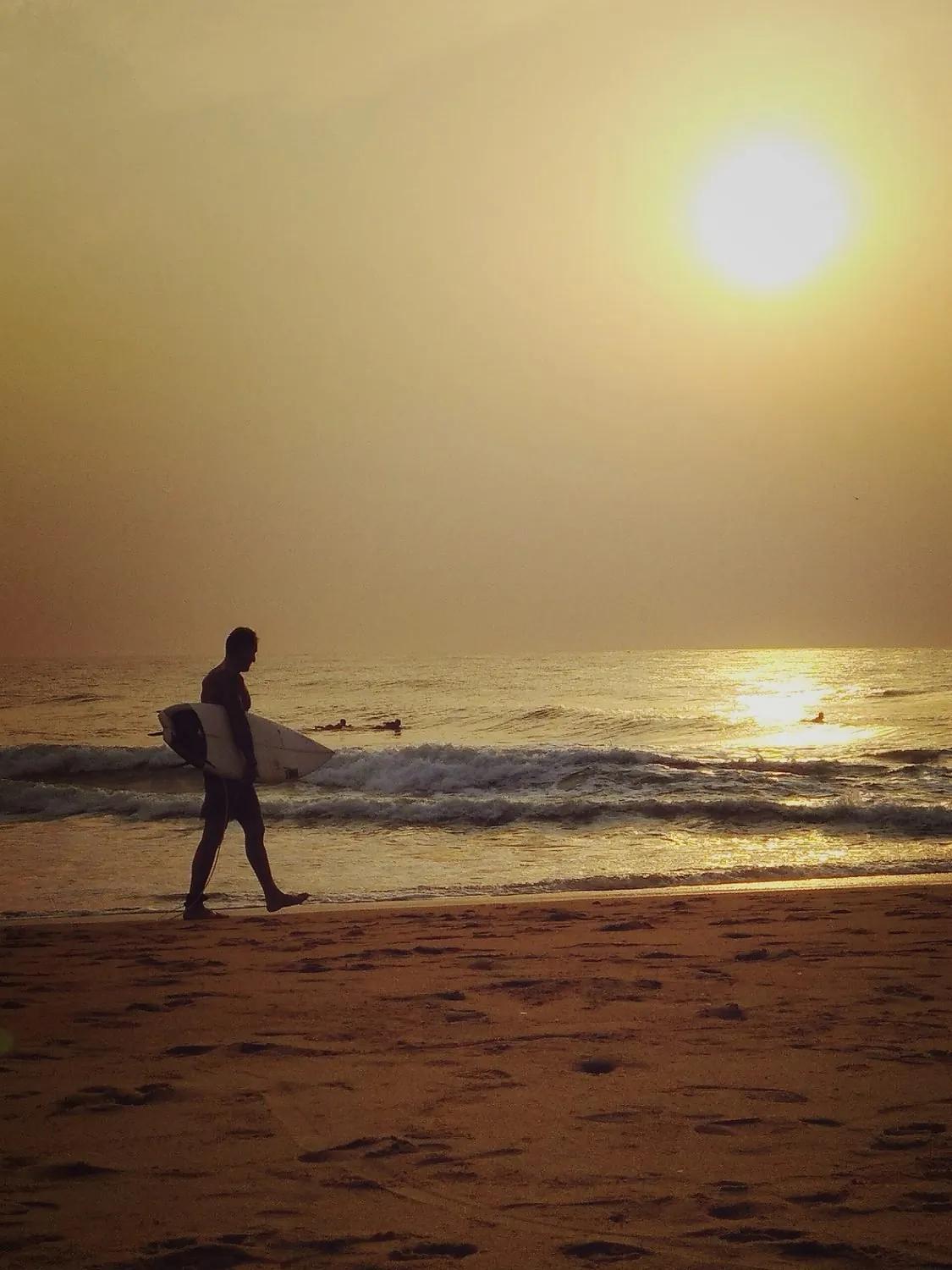 A surfer in silhouette walking along a beach at golden sunset carrying a surfboard, small waves breaking behind