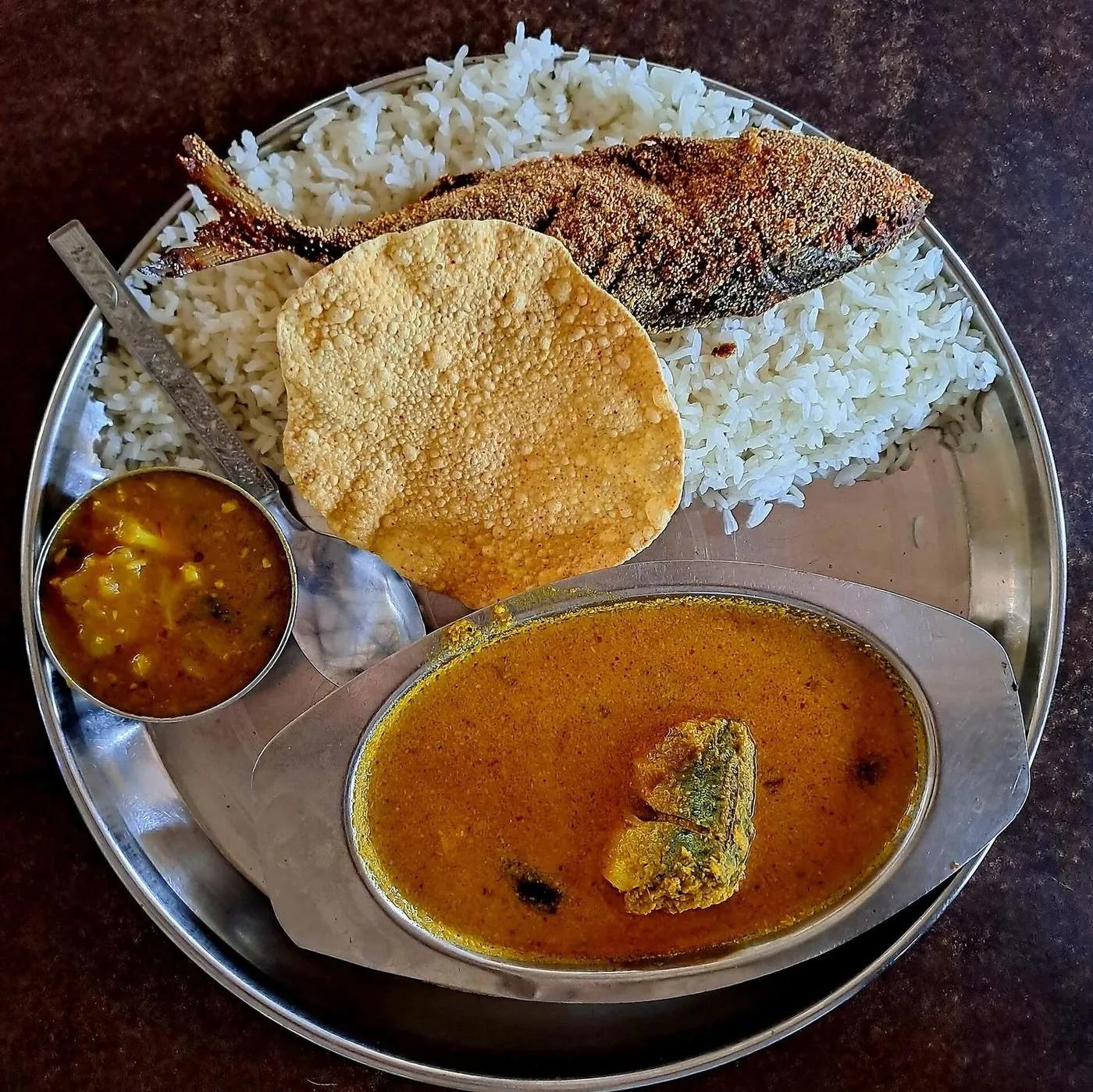 A Goan fish thali on a steel plate: white rice, a whole fried fish, a papad, a bowl of orange coconut fish curry and a small side