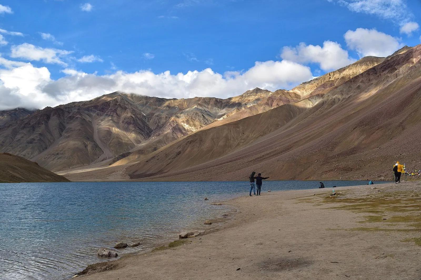Chandratal Lake at 4,300 m in Lahaul-Spiti, turquoise water lapping the gravel beach, rust-and-green ridges of the Chandra Bhaga ranges behind