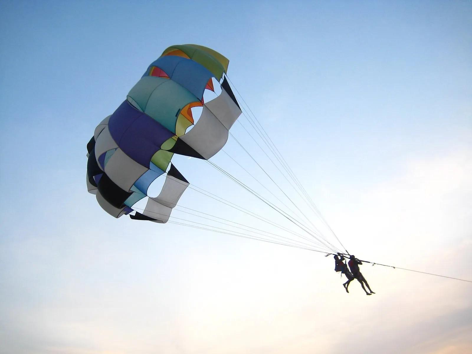 A colourful parasail canopy high over the sea at Calangute beach, Goa, two people in the harness silhouetted against a hazy sky, the tow cable taut and clearly visible