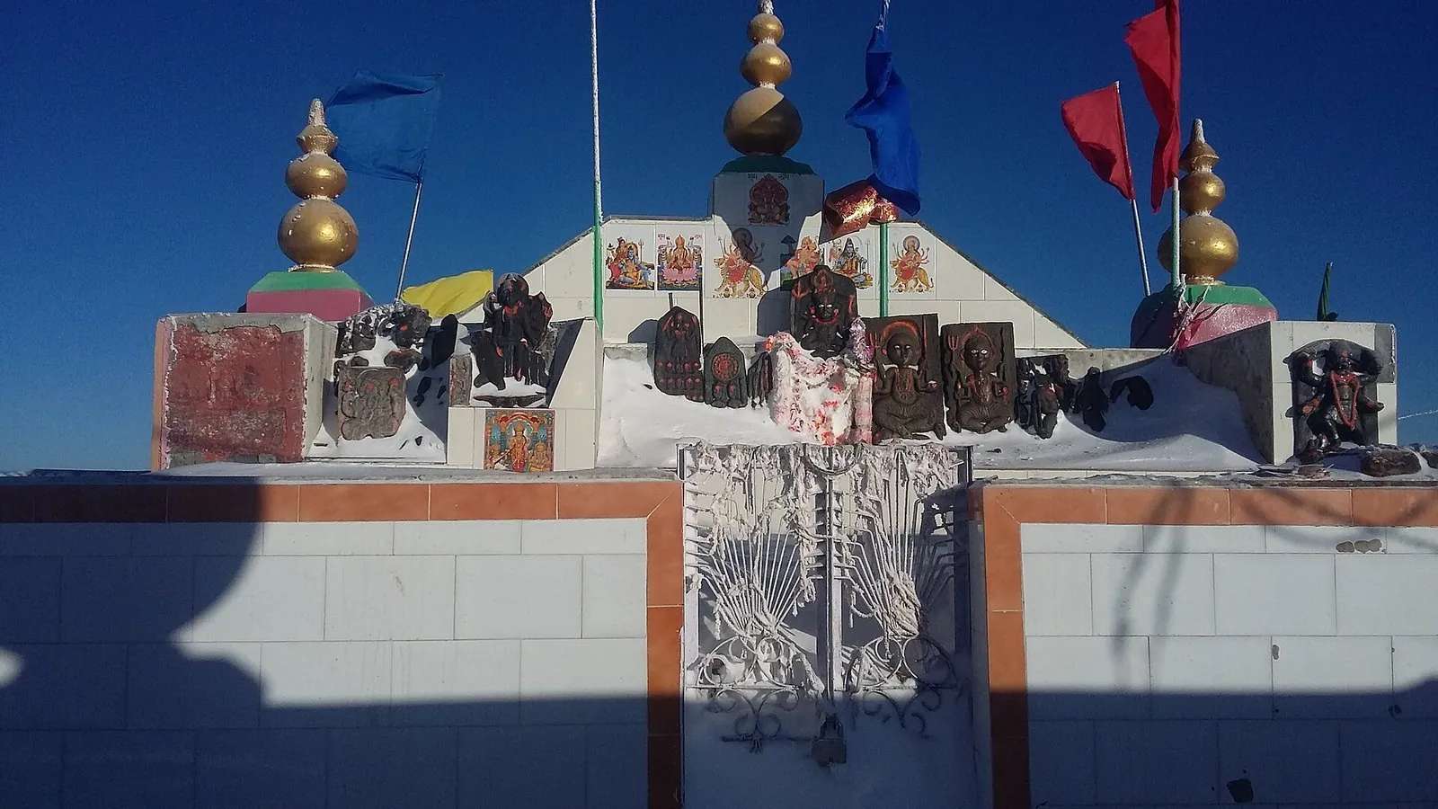 Shikari Devi Temple at 3,359 m in Mandi district, Himachal Pradesh — open-air sanctum with painted deity plaques, gold finials, blue and red prayer flags, and snow still on the wall around the shrine