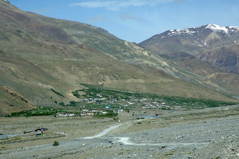 Losar village seen from across the valley. A cluster of low whitewashed houses laid out along a flat green river meadow, brown ridges with snow streaks rising behind, dirt road threading through the foreground