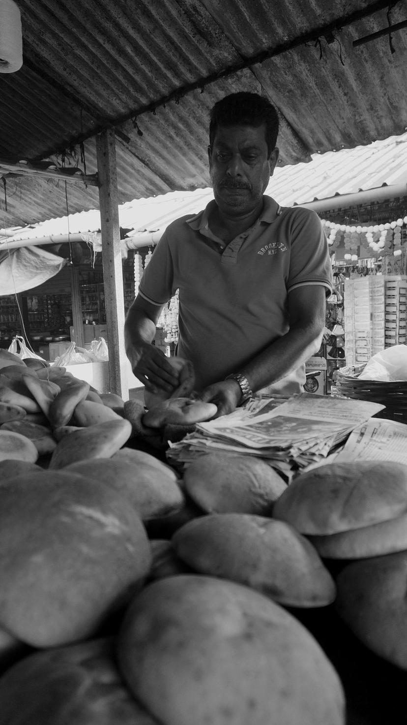 A black and white photo of a Goan bread seller at the Mapusa market behind a mound of round Goan breads, wrapping one, stalls behind