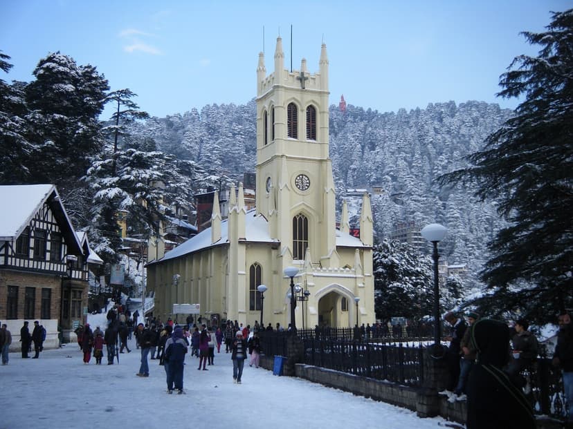 Christ Church Shimla in snow, Neo-Gothic yellow stone tower with clock, snow-covered deodar trees behind, people walking on the cleared square