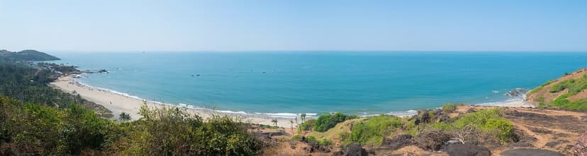 A panorama of Vagator beach in North Goa, turquoise sea, a curved sandy bay and red laterite headlands under a clear sky
