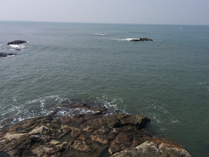 A rocky Goan shoreline with wet foreground rocks in shallow water and small offshore rock outcrops in a calm green-blue sea under a hazy sky.