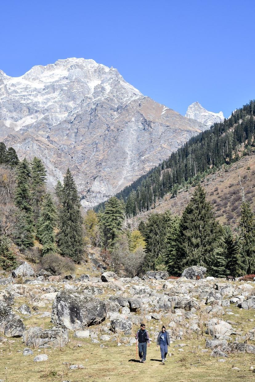 Two hikers crossing a boulder-strewn high pasture toward a snow peak on the Kutla trail, Parvati Valley