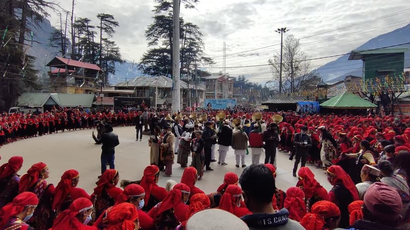 Hundreds of performers in red caps dancing Nati in a circle at the Manali Winter Carnival with deodar trees in the background