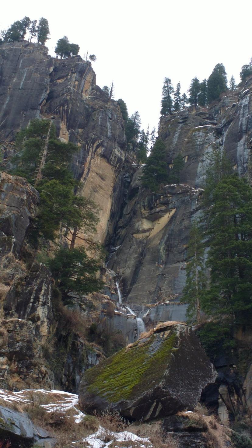 Jogini Falls cascading down a tall rocky cliff face near Vashisht Manali, pine trees clinging to the rock, snow patches on ground