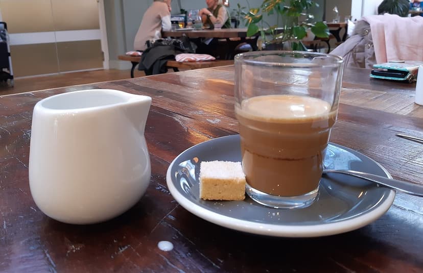 A cortado in a glass on a saucer on a wooden cafe table, other tables behind