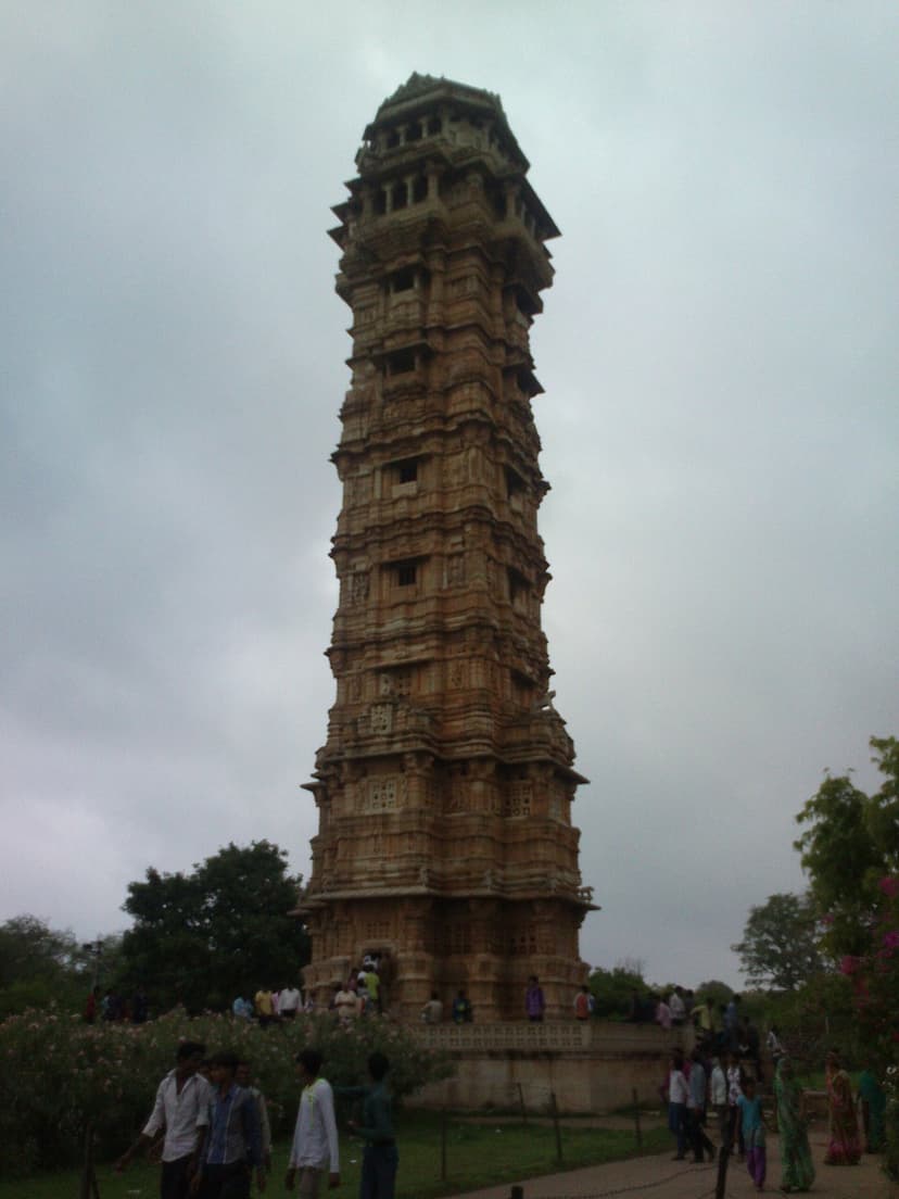 The Vijay Stambh or Tower of Victory, a tall nine-storey carved sandstone tower inside Chittorgarh Fort, under a cloudy monsoon sky with visitors at its base.