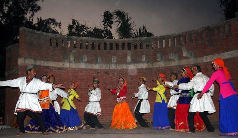 Sirmauri Nati troupe in a line at night against an amphitheatre wall, performers in white tunics and brightly coloured ghagra skirts mid-step, fast choreography