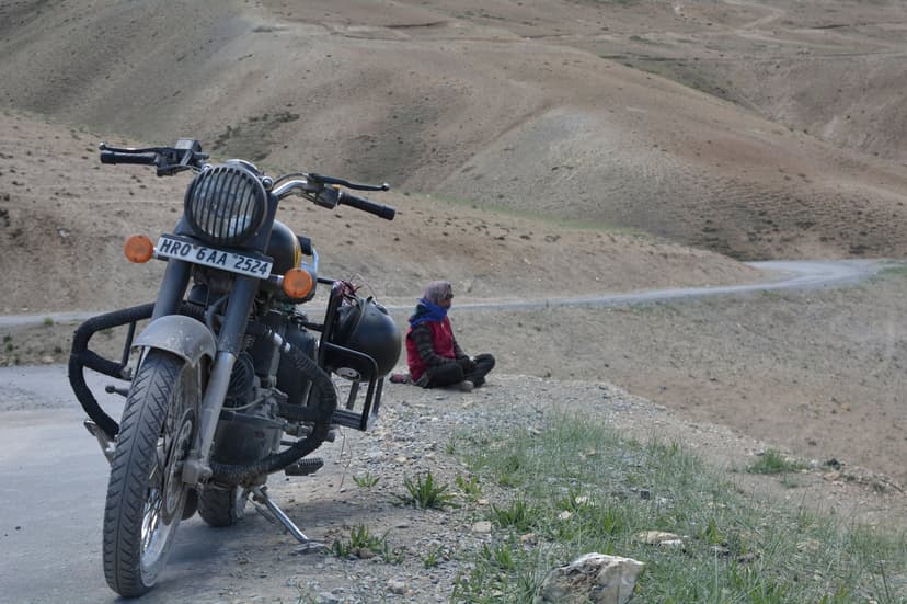 Black Royal Enfield Classic motorcycle parked on a narrow paved road in Spiti with a rider in a red jacket sitting cross-legged on the grass verge, facing away from the camera, looking across brown rolling hills