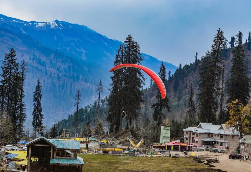 A red paraglider coming in to land at Solang Valley near Manali with deodar forest, activity huts and snow peaks behind