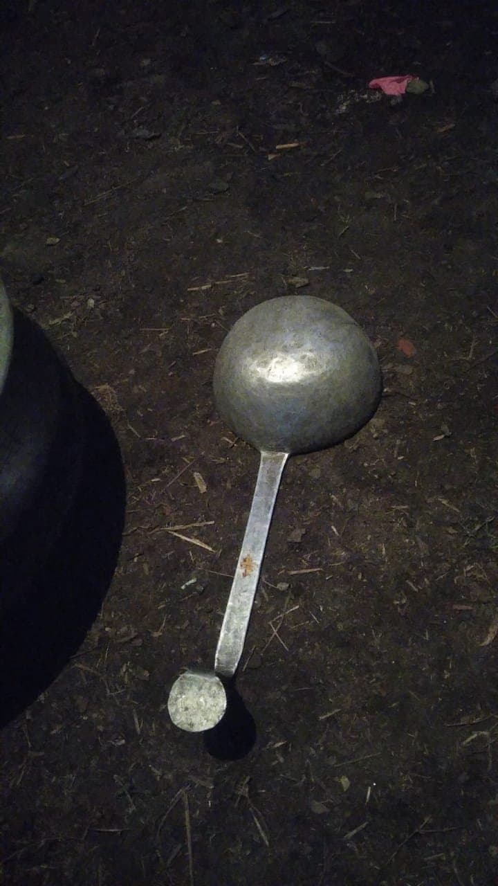 A large hand-hammered metal serving ladle (karchhi) lying on dirt ground at night — the long-handled serving spoon a Boti's team uses to ladle dal and madra from a charoti vessel into rows of waiting pattal plates at a Dham