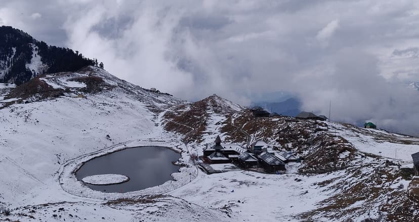 Prashar Lake under snow in winter, with the floating vegetation island still visible as a darker oval inside the lake, and the pagoda temple roof carrying a thick crust of snow