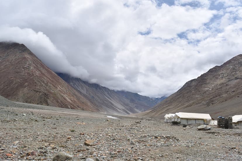 Wide grey gravel apron at Batal in Lahaul, low cloud spilling down the side ridge, a row of white tin-roofed dhabas and tents on the right, brown and rust mountains to the left under a heavy summer sky