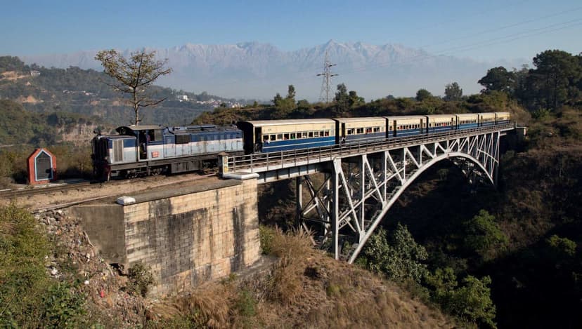 A narrow-gauge train on a long steel arch bridge over the Reond Nullah gorge with the Dhauladhar range in the distance