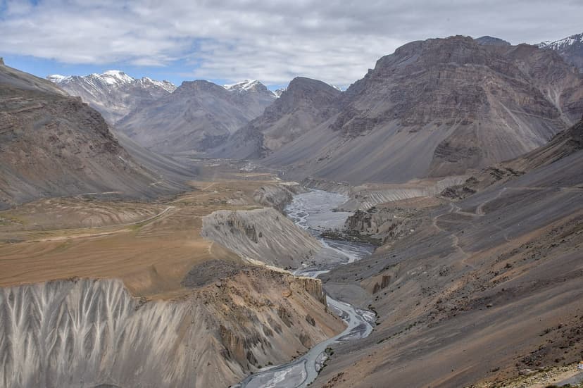 A wide aerial-style view of the upper Spiti valley between Kaza and Losar, narrow river running through pale grey gravel banks below, snow peaks on the far left, the brown-orange terrace above the erosion wall, exactly the corridor Negi's eastern route follows up to Kunzum