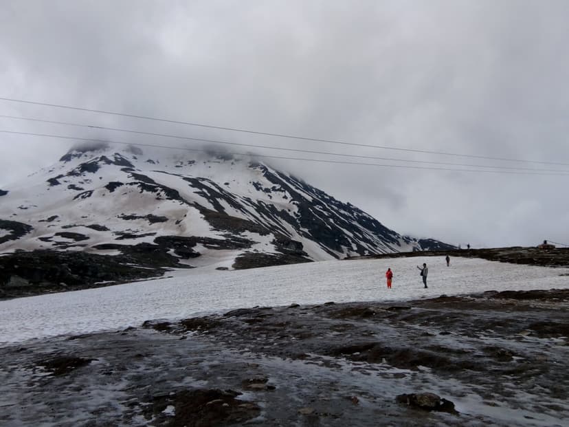 Snow still covering the top of Rohtang Pass under a heavy overcast sky, with a few tourists standing on packed snow and a bare rocky ridge behind