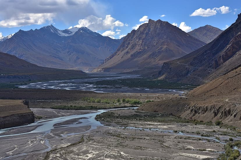 Wide flood plain of the Spiti river. Pale braided channels running through grey gravel, brown ridges on either side and snow-streaked peaks at the head of the valley, dry shrubs in the foreground