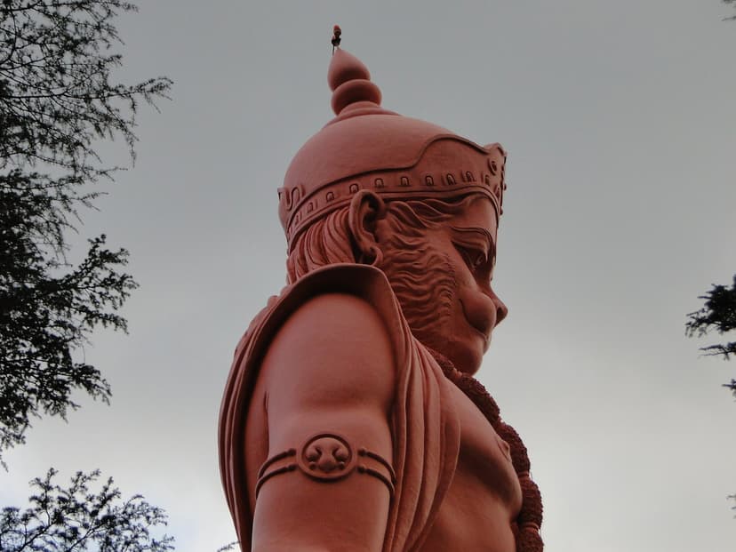 Close-up of the massive 108-foot orange Hanuman statue at Jakhoo Temple Shimla, shot from below against an overcast sky, deodar branches framing the edges