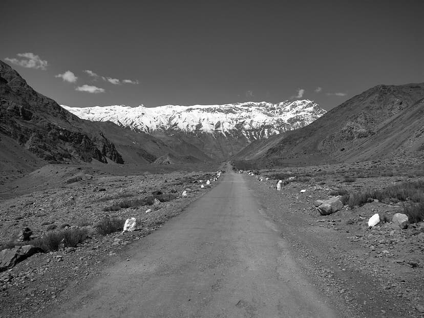 Black-and-white shot of an empty single-lane road running straight into a snow-capped Himalayan ridge, painted boulders marking the verges, a single small figure walking the centreline far ahead