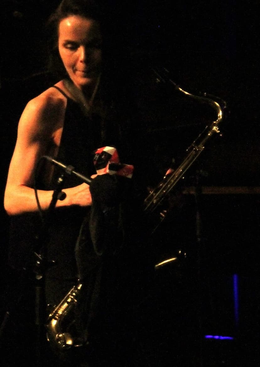A saxophonist on a dark stage in warm light, holding two saxophones, absorbed in playing, no venue signage