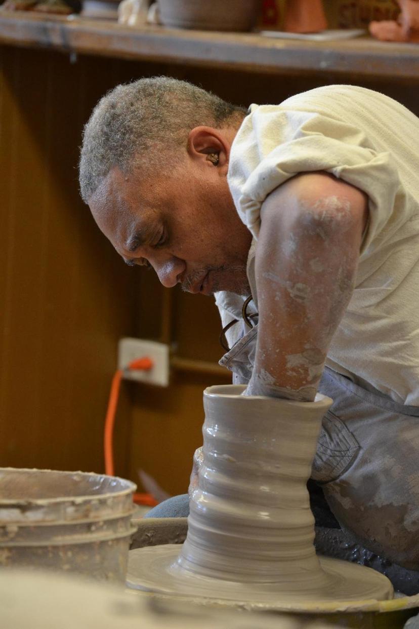 A potter with clay-covered arms throwing a tall grey pot on a wheel