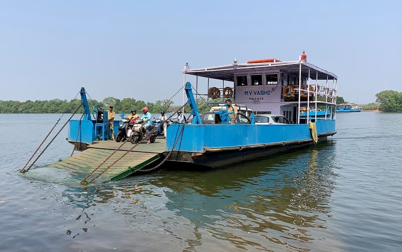 A blue flat-bottom vehicle ferry loaded with motorbikes and cars, ramp down, crossing the Mandovi river with green mangrove banks behind