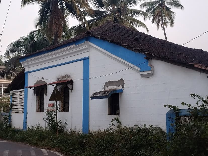 A whitewashed Portuguese-era Goan village house with blue-painted trim and a tiled roof beside a quiet road in Parra, North Goa, coconut palms behind.