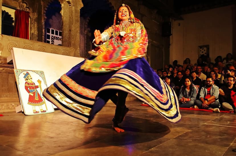 A Rajasthani folk dancer in a bright colourful ghagra spinning mid-performance at the Dharohar dance show at Bagore Ki Haveli in Udaipur, a seated audience watching and a miniature painting on an easel beside her