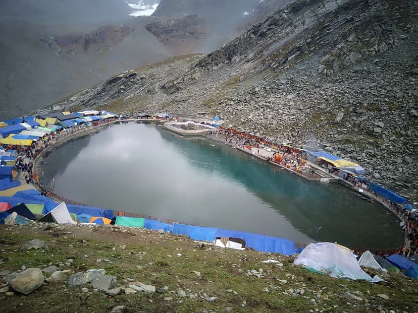 Elevated view of the lake and tent encampment at the Manimahesh Yatra — hundreds of colourful tarps and blue tents wrap the lakeshore and the surrounding boulder slopes