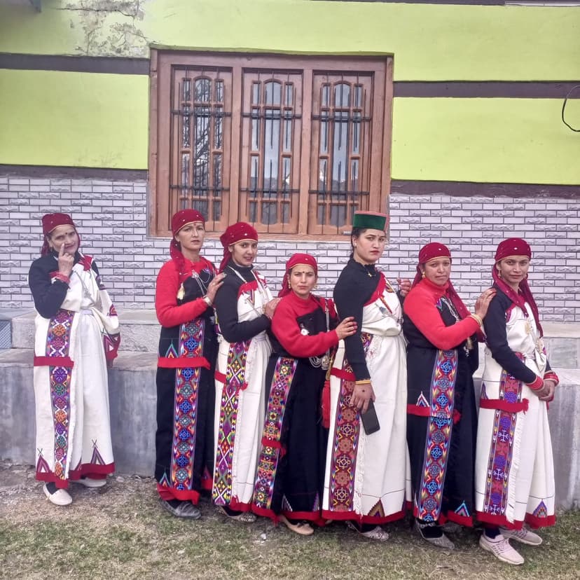 Six women of a Kullu Mahila Mandal standing in a row in traditional white pattu shawls with red and black geometric borders, red dhattu caps, embroidered diamond-pattern skirts, one man in a black topi standing among them