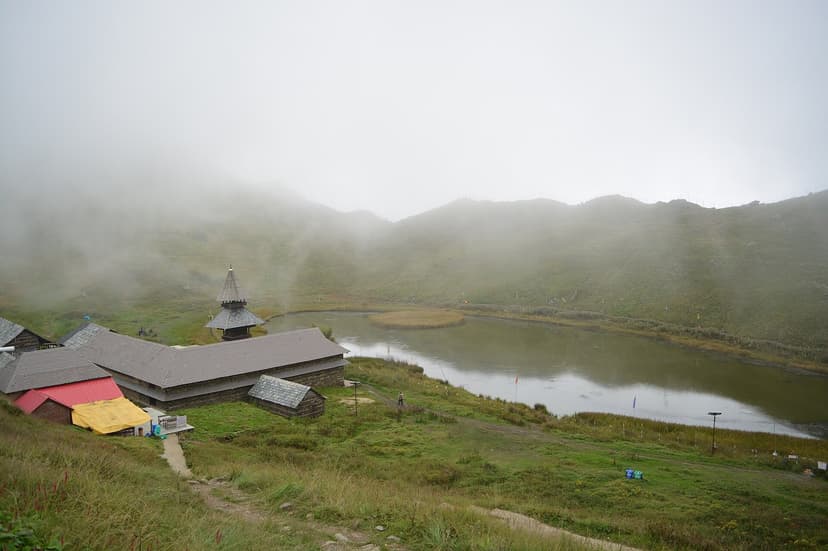 Prashar Lake under low cloud at 2,730 m, with the conical wooden pagoda temple beside the grass-fringed water