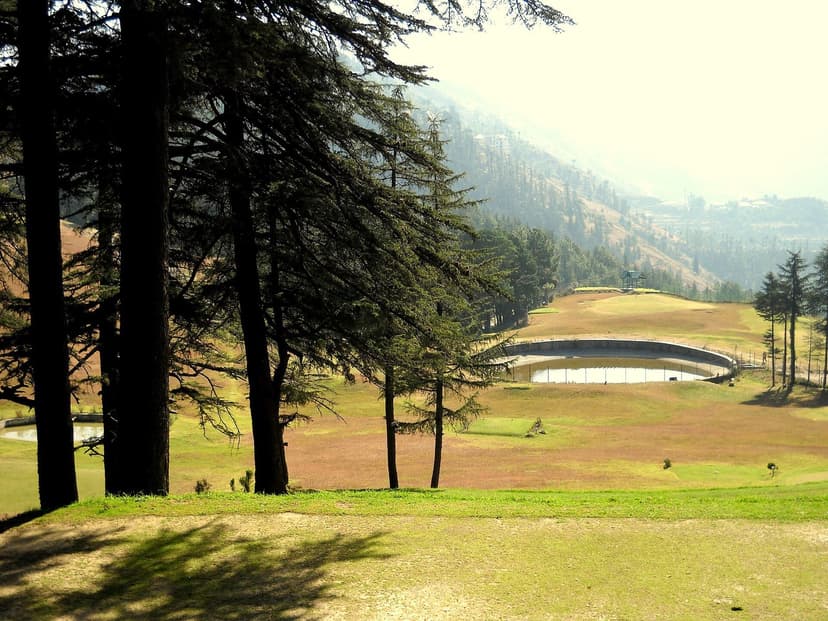 Naldehra Golf Course fairway with tall cedar and deodar trees, misty green hills in the background, a water feature on the course