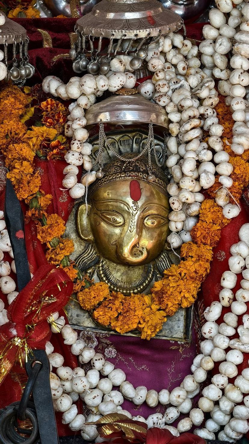 Close-up of a brass deity mohra (metal mask face) with red tilak, draped in white beads and marigold